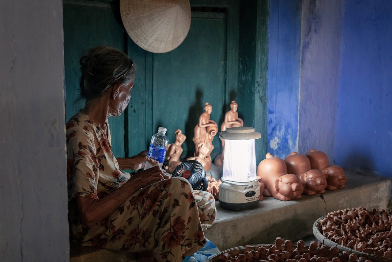 An elderly woman shapes clay figurines illuminated by lamp in a traditional Vietnamese setting.