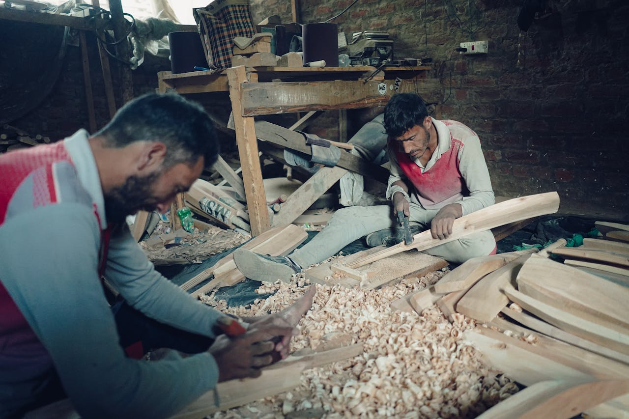 Two craftsmen skillfully carve wood in a rustic workshop setting, surrounded by wooden shavings and tools.